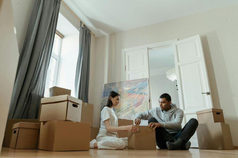Two people unpacking moving boxes on the floor of a bright room in a new home.