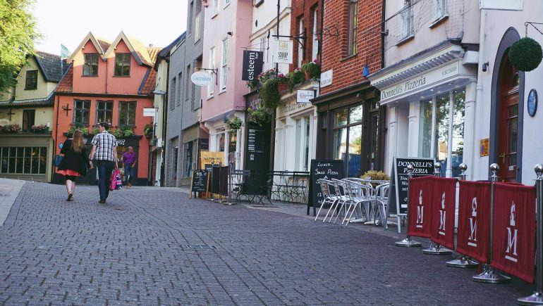 Pedestrians walking along a quiet street with shops and outdoor seating in a compact town center.